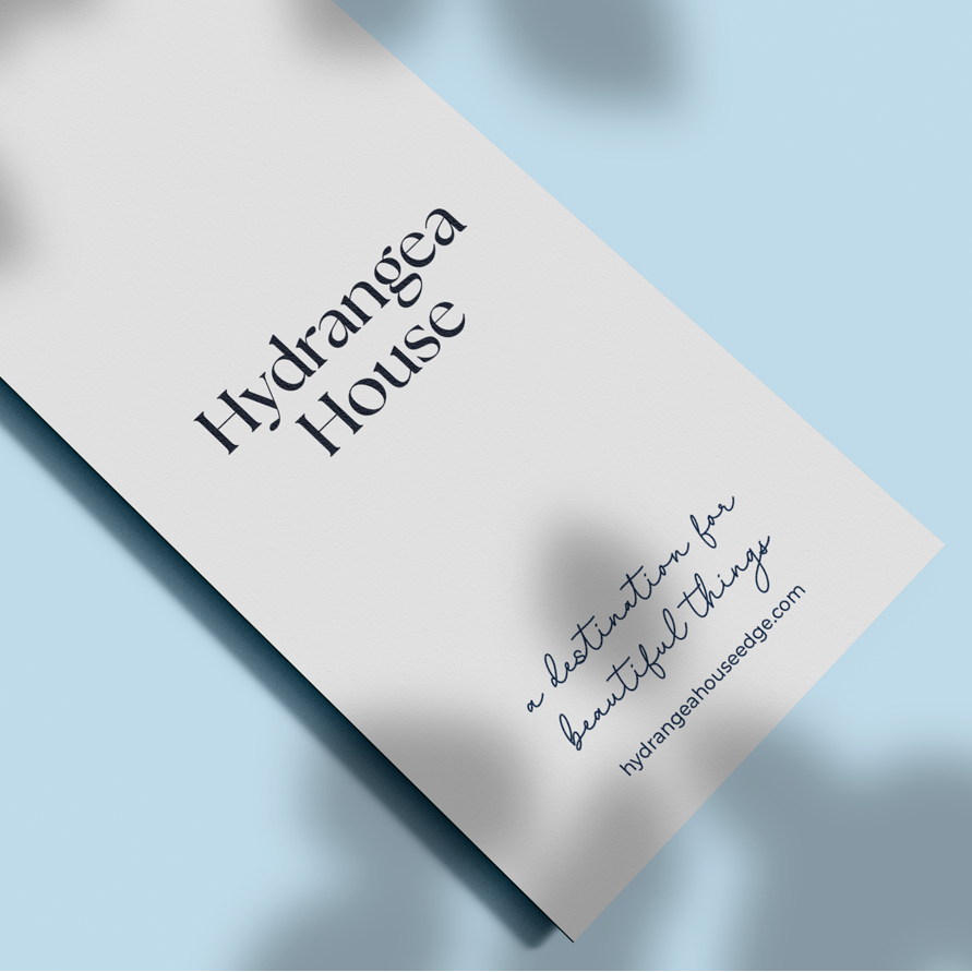 A white paper hang tag with the text 'Hydrangea House' and smaller script text below, attached to a thin string against a light blue background with soft leaf shadows.